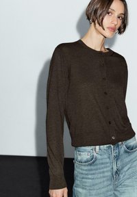 Woman with short brown hair wearing a dark brown buttoned cardigan and light blue high-waisted jeans against a plain background.