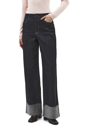 Kvinde iført sorte jeans med høj talje og vide ben med frynser ved kanten, sorte hælede sko og en lyserød top med lange ærmer, hånd i lommen.