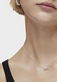 Collar de cadena de plata con tres pequeñas piedras preciosas brillantes. Lleva puesto en un cuello suave, visto contra un fondo de ropa oscura.