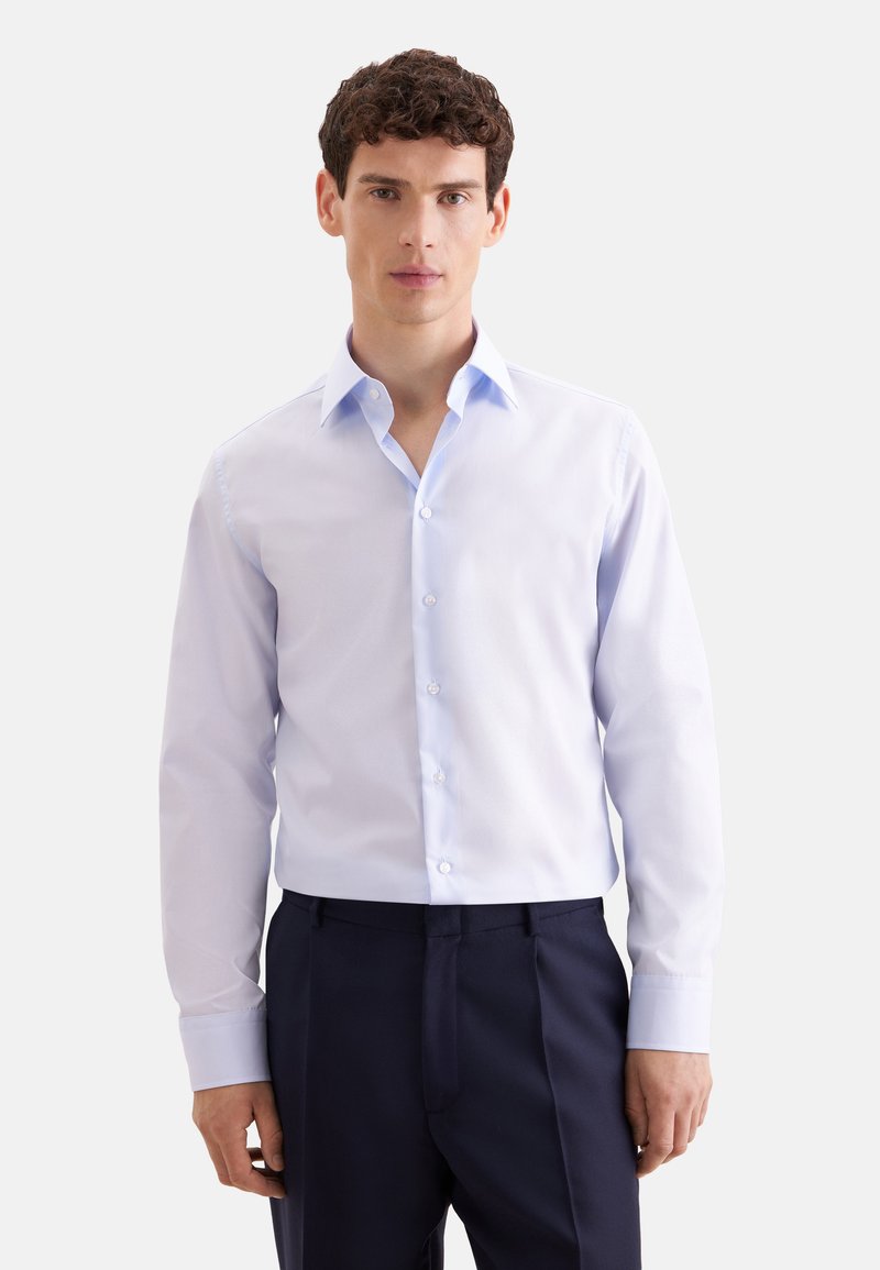 Jeune homme aux cheveux courts et bouclés portant une chemise bleu clair boutonnée et un pantalon foncé, debout devant un fond clair uni.