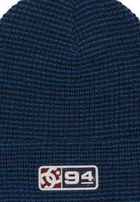 Bonnet en tricot bleu foncé avec un motif texturé. Présente un patch logo en rouge, blanc et marine avec le design "94".
