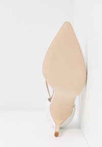 Chaussure à talon haut blanche avec un bout pointu et une surface texturée, dotée d'une fine bride de cheville et d'une semelle extérieure couleur beige avec une légère adhérence en caoutchouc.