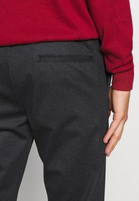 Pantalon gris à carreaux avec une texture lisse et une poche arrière. Un pull rouge est partiellement visible au-dessus de la taille.