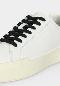Λευκό sneaker χαμηλού προφίλ με μαύρα κορδόνια και εκρού σόλα, λείο δερμάτινο επάνω μέρος, διακριτικό ανάγλυφο λογότυπο στο πλάι.