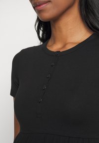 Primer plano de una mujer que lleva una camiseta negra de manga corta con un escote redondo y una fila vertical de seis botones negros en el pecho.