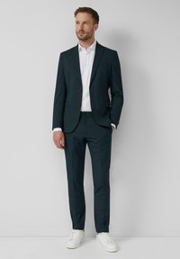 Costume bleu canard avec blazer à deux boutons et pantalon assorti, associé à une chemise blanche et des baskets blanches. Tissu lisse, coupe ajustée.
