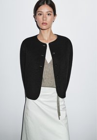 Kvinna med mörkt hår bär en svart knappad cardigan över en beige topp och en vit kjol, stående mot en enhetlig ljus bakgrund.