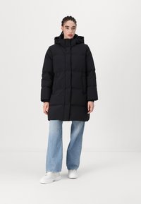 Black puffer coat with a hood, featuring horizontal quilted sections, front pockets, and long sleeves, paired with light denim and white sneakers.