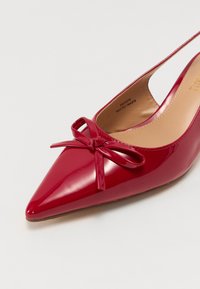 Scarpa slingback in vernice rossa con punta appuntita, finitura lucida e piccolo motivo a fiocco sulla parte anteriore. L'interno presenta una fodera beige.
