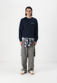 Marineblauer Sweatshirt mit dem Text "Club 1990", graue Nadelstreifen-Hosen mit weitem Bein und ein blaues Karoshirt, das um die Taille gebunden ist. Weiße Turnschuhe runden den Look ab.