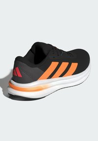 Zwarte sportieve sneaker met oranje accenten en drie zwarte strepen, met een gestructureerde bovenkant en een witte Cloudfoam-zool voor demping.