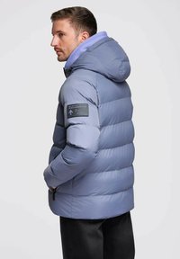 Jasnoszara pikowana kurtka z kapturem, z fioletową podszewką i czarną naszywką na lewym rękawie, kieszeniami na zamek i fakturowaną powierzchnią.