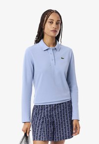 Polo a maniche lunghe di colore azzurro chiaro con colletto a bottone e logo verde. Abbinato a pantaloni corti navy con motivi geometrici.
