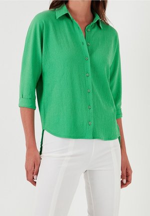Femme portant un chemisier vert vif à manches longues boutonné avec un pantalon blanc sur un fond neutre.