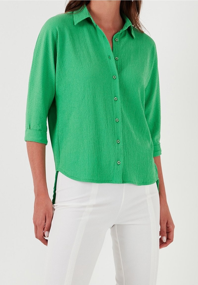 Femme portant un chemisier vert vif à manches longues boutonné avec un pantalon blanc sur un fond neutre.