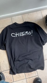 Crni majica položen na pločicama sa natpisom "CHEERS!" ispisanim velikim bijelim slovima na leđima.