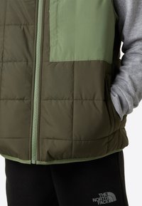 Vadderad väst i mörkgrön med en ljusgrön bröstpanel. Har dragkedjestängning och sidofickor, tillsammans med en grå sweatshirt.