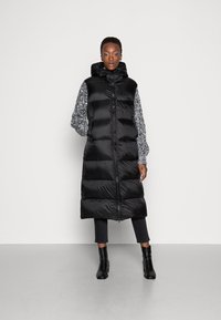 Gilet imbottito nero con cappuccio, caratterizzato da una finitura lucida, design a trapuntatura e maniche grigie testurizzate. Si chiude con una zip centrale.