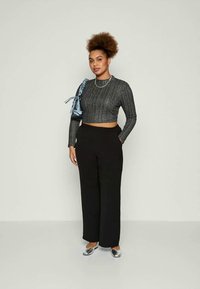 Femme aux cheveux bouclés portant un crop top à côtes gris, un pantalon large noir, des ballerines argentées, un collier en chaîne et portant un petit sac à bandoulière bleu.