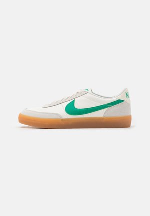Biały niski sneaker z zielonym logo Nike i gumową podeszwą w kolorze gumy, widok z boku na jasnym, gładkim tle.
