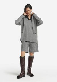 Grauer übergroßer Pullover mit geripptem Saum, graue Kabelstrick-Shorts und glänzende bordeauxfarbene kniehohe Stiefel. Texturiertes Strickdesign bemerkt.