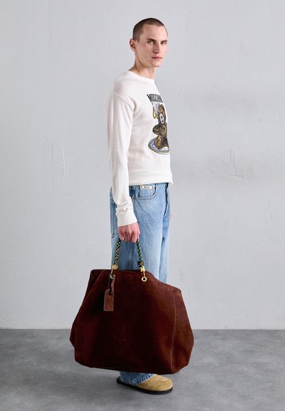 Geantă de tote din suede maron cu o formă lejeră și mânere împletite colorate. Pură de un model îmbrăcat într-un pulover grafic alb și blugi albaștri deschisi.
