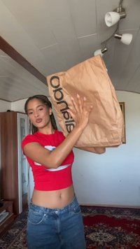 Ruskea paperikauppakassi mustalla logolla, jonka yläosa on avoin. Henkilöllä on punainen paita ja siniset farkut, ja hän pitää kassia ylhäällä.