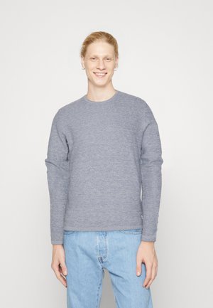 Maglione grigio a maglia testurizzata con maniche lunghe e collo rotondo, abbinato a jeans azzurri chiari. Il design è minimalista e casual.