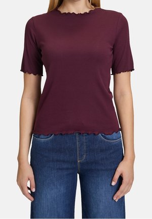 Vrouw draagt een aansluitend, bordeauxrood geribd topje met korte mouwen en gegolfde randen, gecombineerd met een hooggesloten blauwe spijkerbroek.