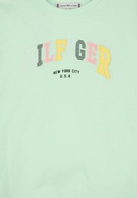 T-shirt in cotone verde chiaro con testo multicolore: "HILFIGER" in rosa, nero e giallo, con "NEW YORK CITY U.S.A." sotto in nero.