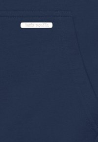 Étiquette en métal argenté avec le texte "bazix republiq" sur un tissu bleu marine près d'une couture de poche cousue.