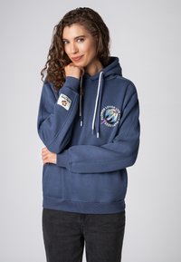 Marineblauwe hoodie met een trekkoord, voorzien van een bloemenafbeelding en logopatches op de mouwen. Zachte stof en een ontspannen pasvorm.