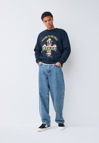 Marinblå sweatshirt med grafik som föreställer skallar i tomteluva. Matchad med ljusblåa baggyjeans och svarta sneakers med vita detaljer.