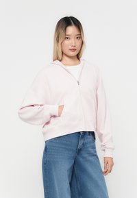 Mlada žena s ravnom, kosom do ramena, obučena u svijetlu ružičastu hoodie s patent-zatvaračem i plave wide-leg traperice, stoji naslonjena na bijelu pozadinu.