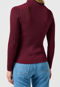 Bordowy sweter prążkowany z wysokim kołnierzem i długimi rękawami, o dopasowanym kroju i lekkim rozciągnięciu. Połączony z niebieskimi jeansami.
