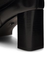 Must mustvalge nahast kõrged kontsad, millel on turske ruudukujuline konts, sile tekstuur ja nähtavad õmblused kontsa lähedal, millel on metallist logo detail.