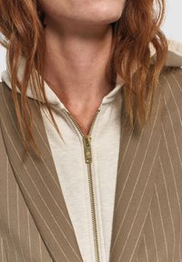 Blazer marrone a righe sottili indossato sopra una felpa crema con cappuccio, caratterizzato da una zip dorata e una texture morbida. Focalizzati sui dettagli del colletto e del cappuccio.