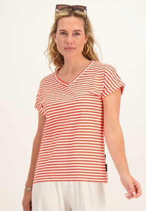 Vrouw draagt een rood-wit gestreept shirt met korte mouwen, witte broek, gouden hoepeloorbellen, zonnebril op het hoofd, staand tegen een effen achtergrond.