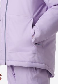 Ανοιχτό μωβ μπουφάν puffer με φερμουάρ μπροστά, πλαϊνές τσέπες και ελαστικά τελειώματα στα μανίκια. Το ύφασμα έχει λείο υφή με διακριτική λάμψη.