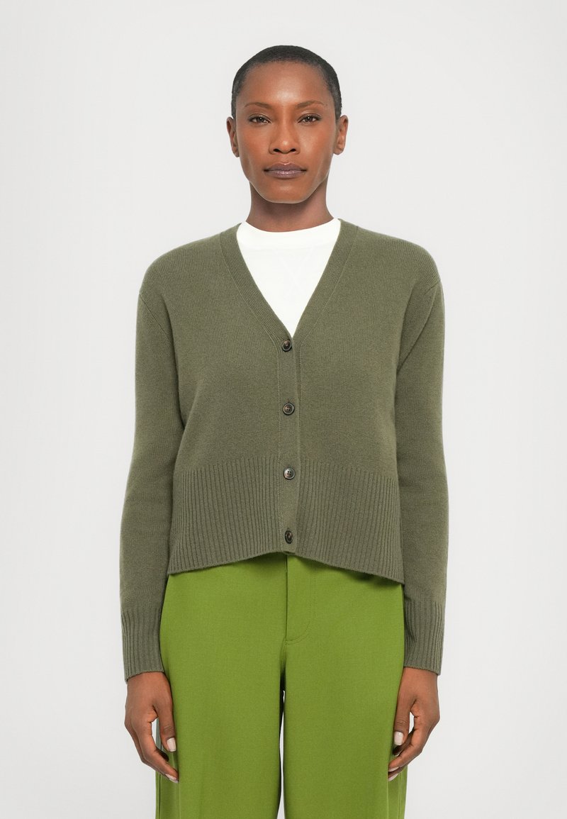 Olive green knitted cardigan with a v-neck, five dark buttons, and ribbed cuffs/hem, paired with bright green pants.
