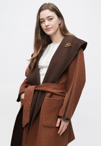 Palton maro și maro închis din amestec de lână, cu guler larg, curea și buzunare mari. Are o broșă aurie și accente cu dungi pe mâneci.