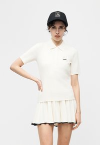 Jeune femme portant un polo blanc à manches courtes et une jupe plissée avec bordure noire, coiffée d'une casquette de baseball noire, posant avec la main sur la hanche.