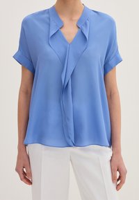 Blusa leggera azzurro chiaro a maniche corte con scollo a V e design frontale drappeggiato. Realizzata in un tessuto morbido e leggero.