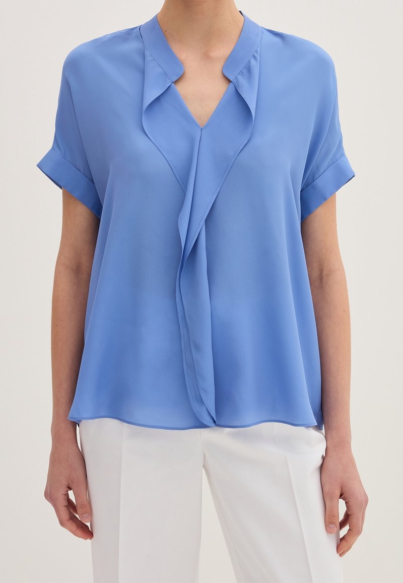 Blusa leggera azzurro chiaro a maniche corte con scollo a V e design frontale drappeggiato. Realizzata in un tessuto morbido e leggero.