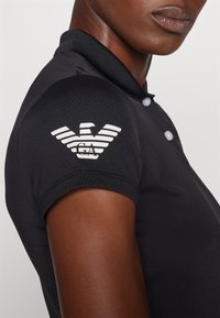 Μαύρο πόλο μπλούζα με ριγωτό γιακά και τρεις κουμπάκια, με λευκό λογότυπο αετού στον αριστερό ώμο, επιδεικνύοντας μια ομαλή, αναπνέουσα υφή.