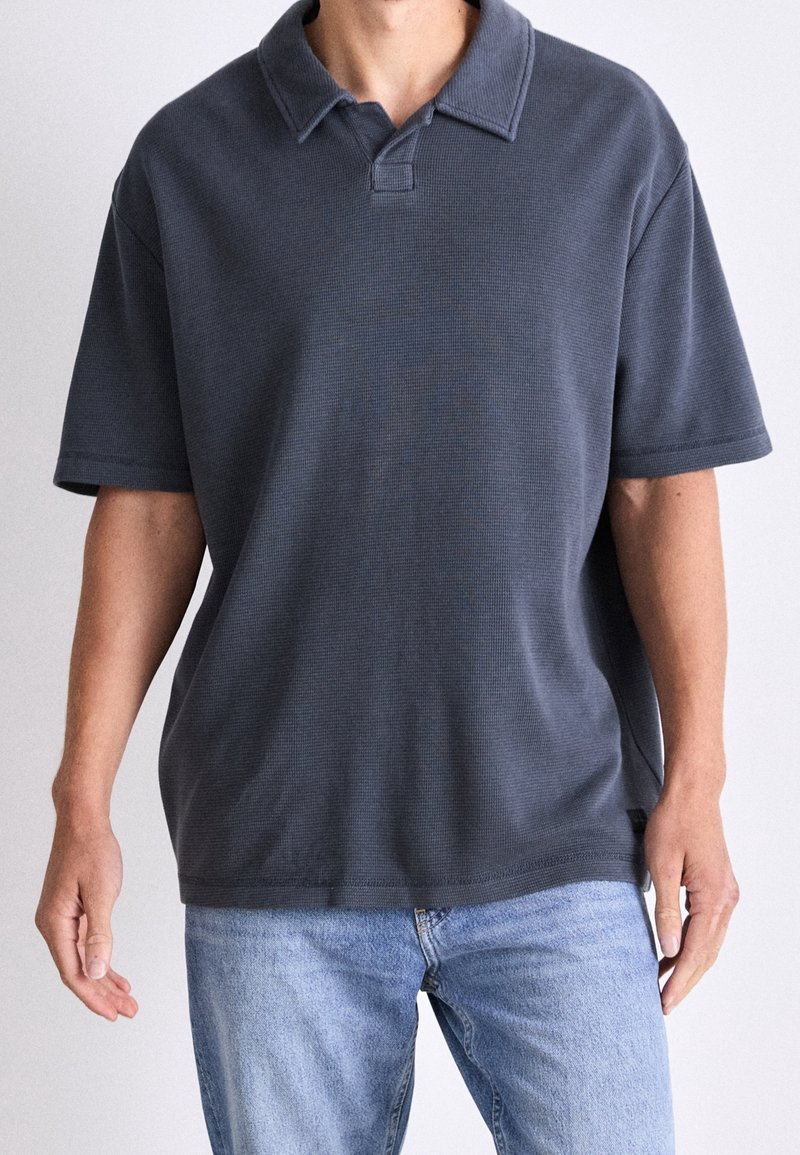 Polo gris foncé à manches courtes avec un col à revers, tissu texturé et coupe décontractée, associé à un jean bleu clair.