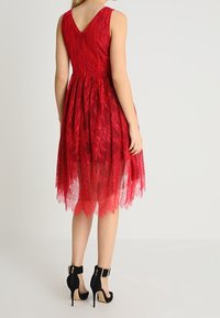 Femme portant une robe rouge en dentelle sans manches avec un dos en V et des chaussures noires à talons hauts avec bride à la cheville, debout sur un fond blanc.