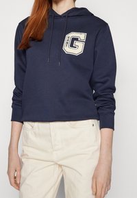 Felpa blu marina con cappuccio e lacci, dotata di un logo "G" ricamato di colore beige chiaro sul petto. Realizzata in tessuto morbido, abbinata a pantaloni color crema.