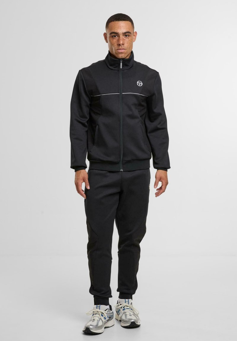 Mann steht in einer schwarzen Zip-Jacke, passenden schwarzen Trainingshosen und weißen Sportschuhen vor einem schlichten weißen Hintergrund.