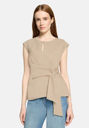 Frau mit glattem roten Haar, die ein beiges ärmelloses Oberteil mit einer Vorderschleife und blaue Jeans vor einem schlichten hellen Hintergrund trägt.